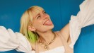 Eine blonde Frau mit schulterlangen blonden Haaren und grünen Haarspitzen schaut nach oben rechts, streckt die Arme in die Luft und streckt die Zunge raus