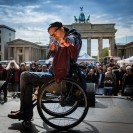 Ein Mann im Rollstuhl auf einer Bühne vor dem Brandenburger Tor zeigt mit den Händen in die Kamera