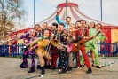 12 Personen in bunten Kostümen mit ihren Instrumenten vor einem Zirkuszelt