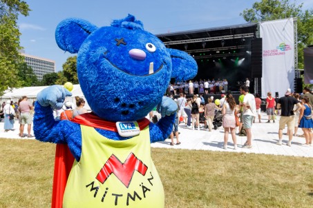 Ein blaues Maskottchen steht vor einer B&uuml;hne und h&auml;lt die Arme hoch