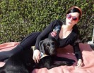 Eine Frau mit langen schwarzen Haaren liegt auf einer fosafarbenen Decke. Sie trägt eine rote Sonnenbrille. Vor ihr liegt ein großer schwarzer Hund.