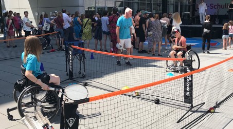 Blick auf ein Rollstuhl-Tennisfeld, auf dem zwei Kinder Tennis spielen