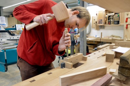 Ein junger Mann in einem roten Pullover bearbeitet mit einem Hammer ein St&uuml;ck Holz.