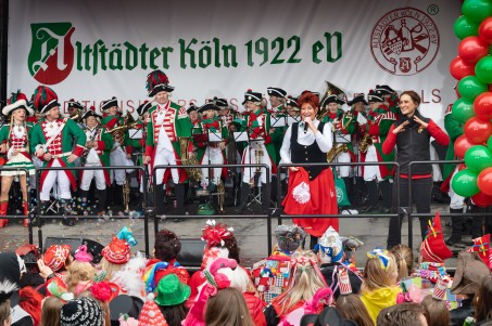 Man sieht ein in traditionelle gr&uuml;n-rot-wei&szlig;e Karnevalsuniform gekleidetes Blasorchester auf der B&uuml;hne bei der Er&ouml;ffnung des K&ouml;lner Stra&szlig;enkarnevals auf dem Alter Markt. Neben einer l&auml;chelnden S&auml;ngerin steht eine Geb&auml;rdendolmetscherin.